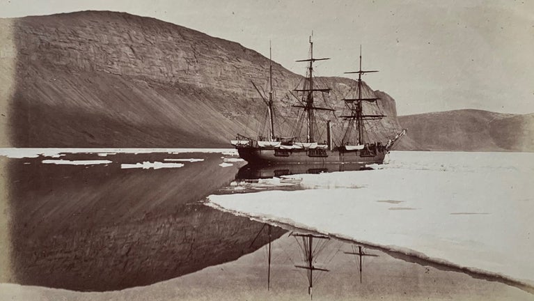 A ship anchored near the coast with a clear reflection in the water and mountains rising on the left-hand side.